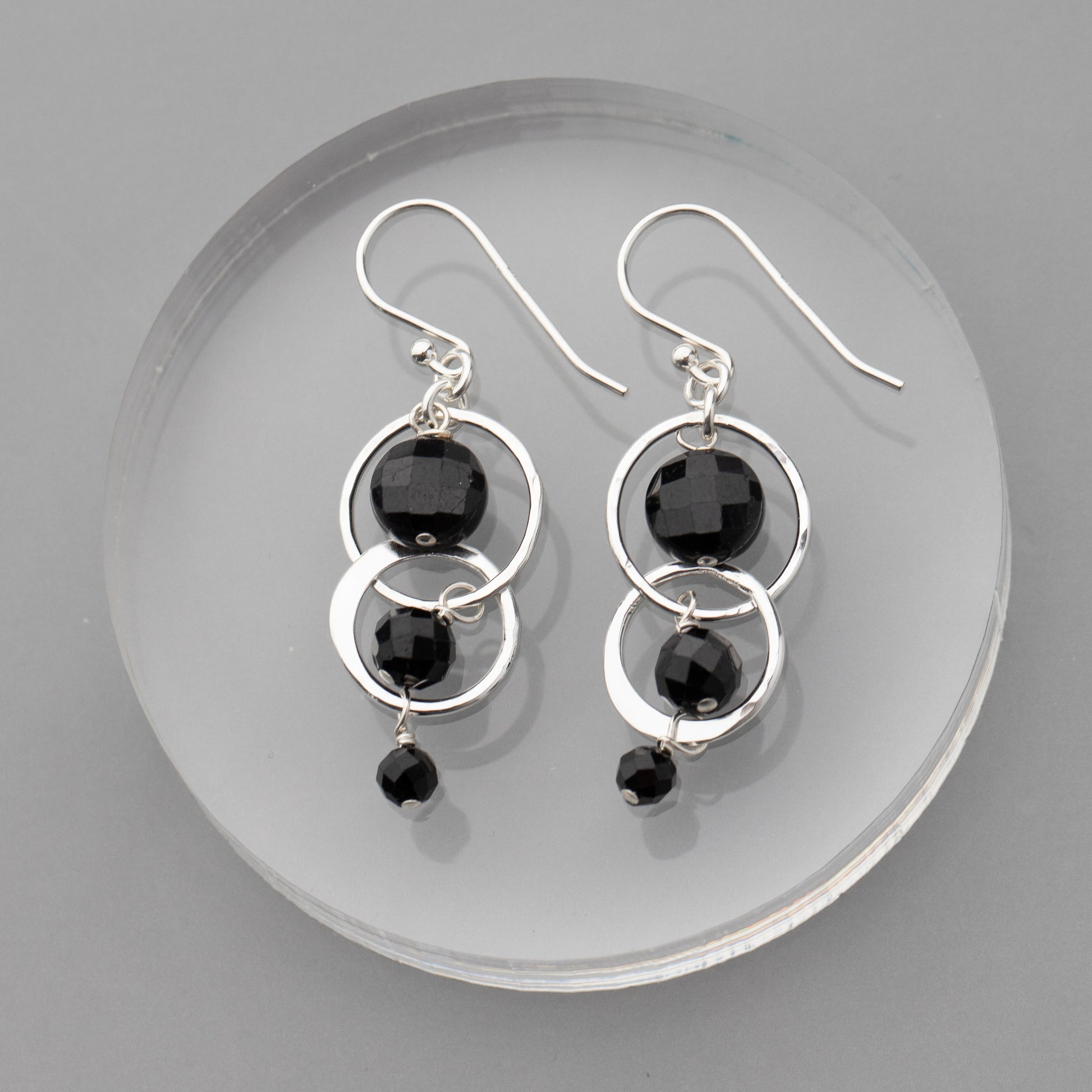 Silver earrings with black tourmaline stones on a clear circular stand against a gray background