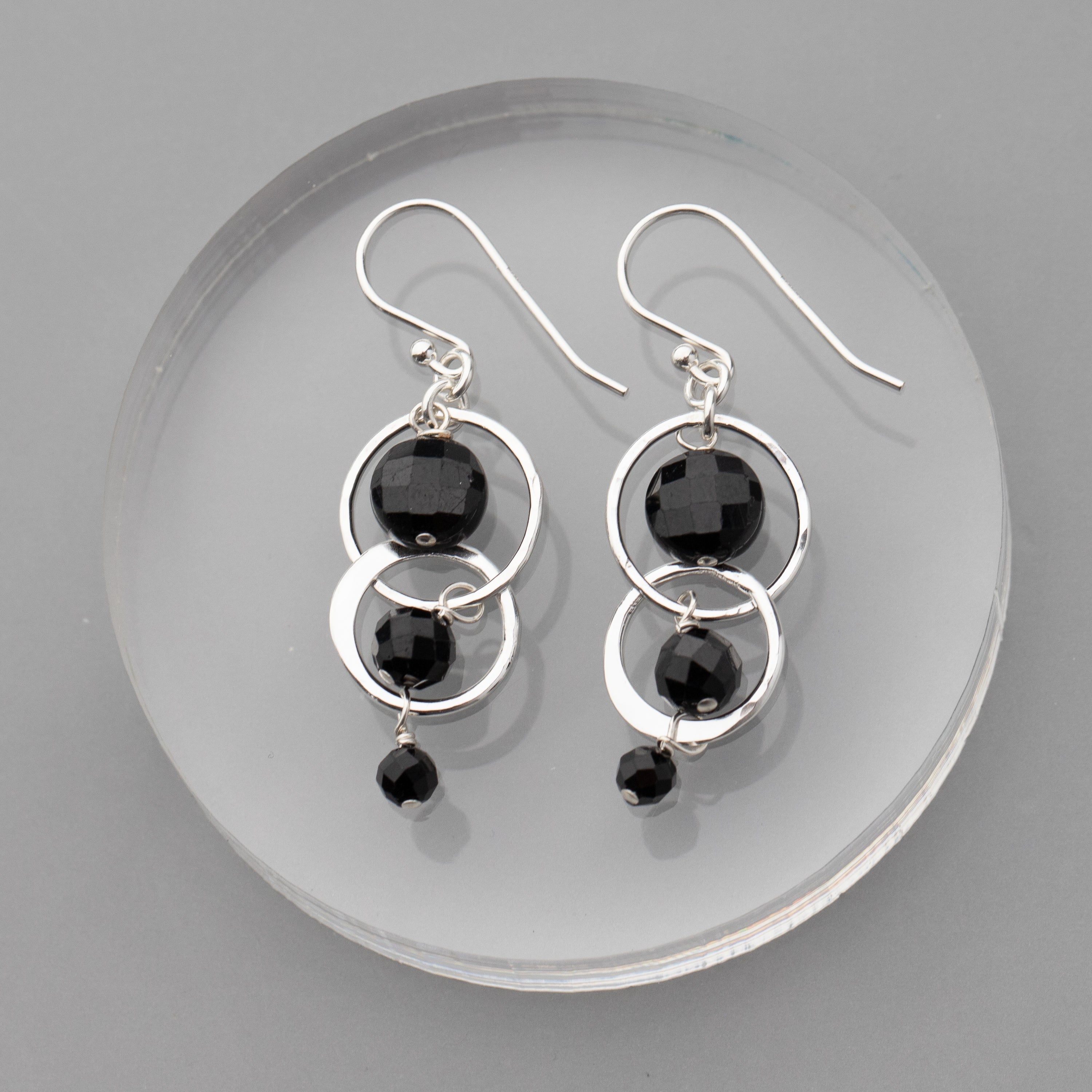 Silver earrings with black tourmaline stones on a clear circular stand against a gray background