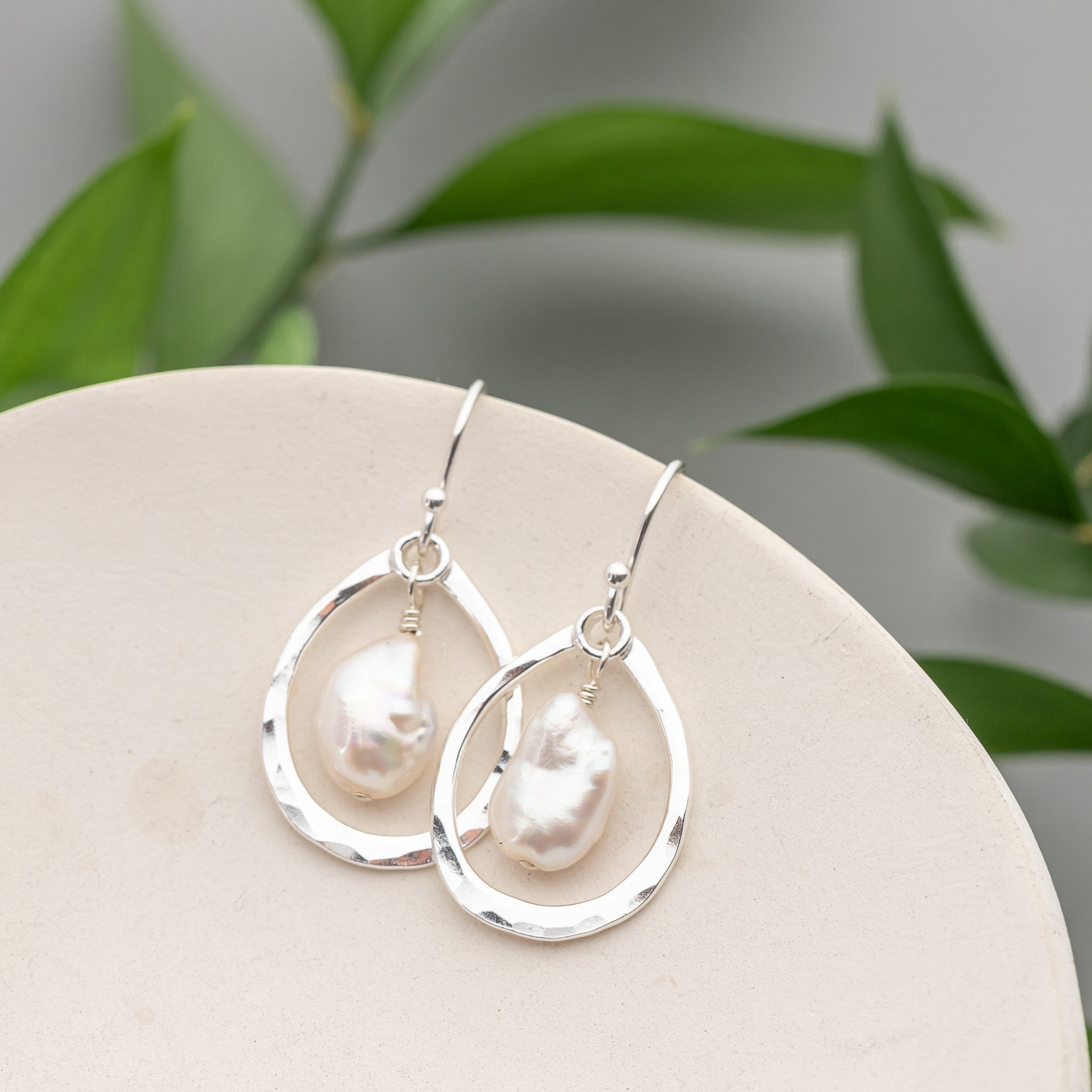 Handmade sterling silver earrings with pearl drops on a white surface with green leaves in the background