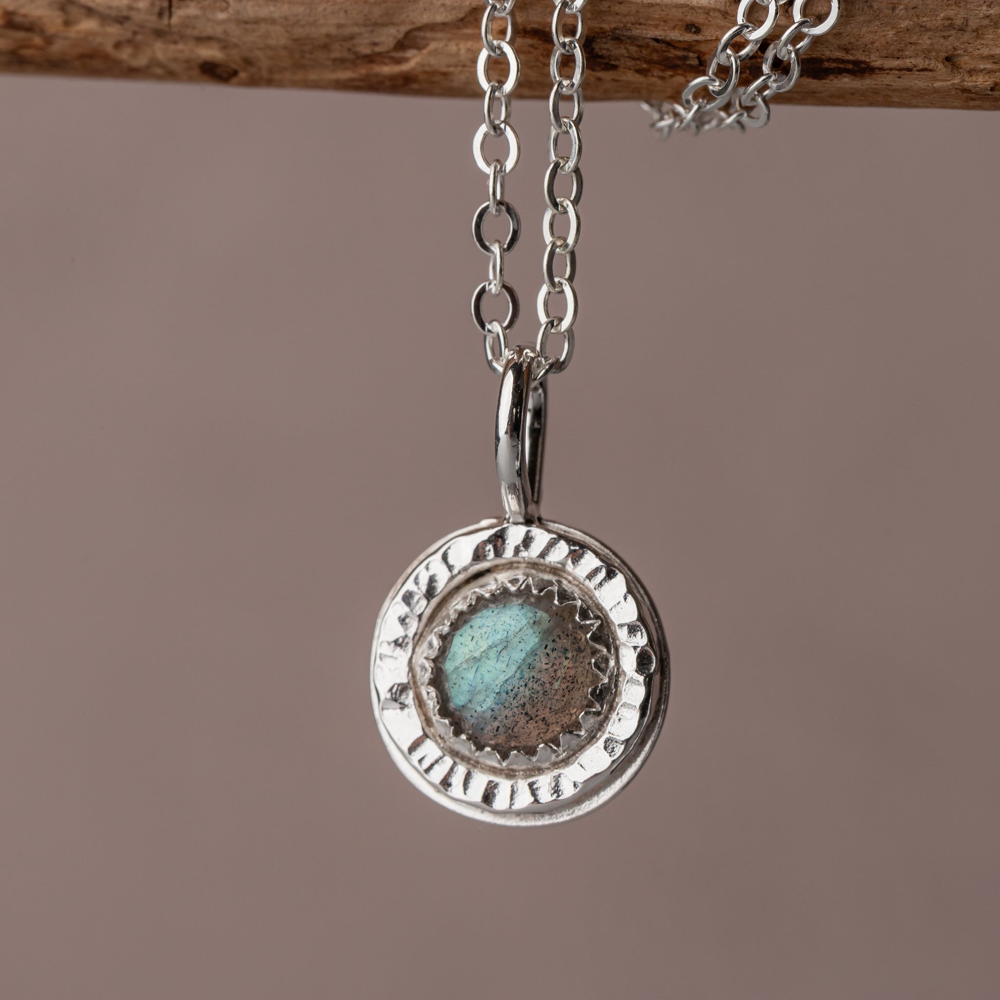 a small labradorite pendant necklace is shown hanging from a branch.  The pendant has a hand hammered frame and a gorgeous blue green flash of color across the surface.
